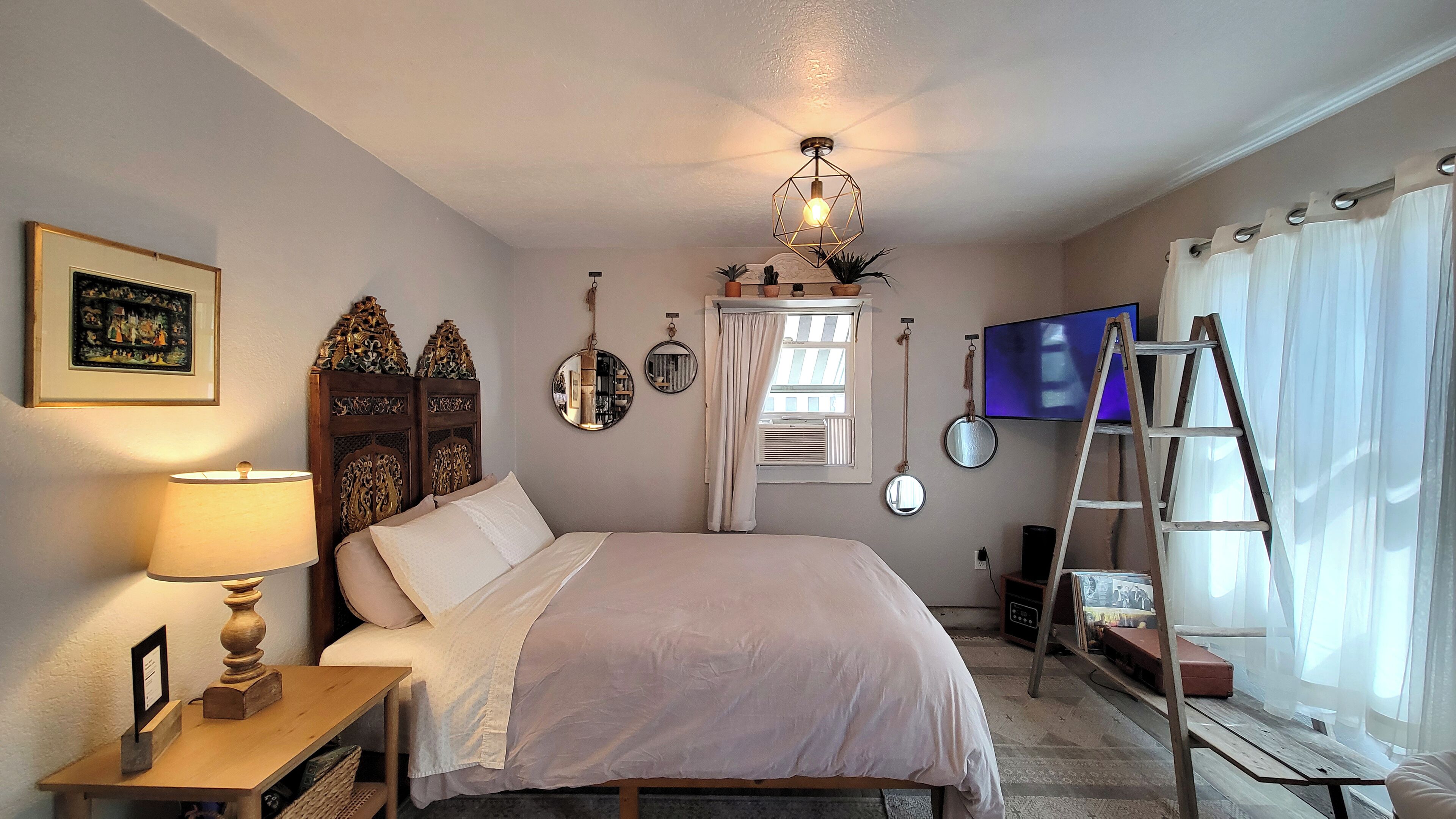 A bedroom with a bed, a wooden nightstand, a lamp, a dresser, a TV, and a ladder.