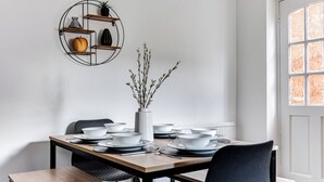 Ruokapöytä on katettu valkoisilla kulhoilla ja lautasilla, pöydällä on maljakko, jossa on oksia, ja seinälle on kiinnitetty hylly, jossa on koriste-esineitä.
