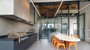 A modern kitchen with a built-in grill, a dining area with orange chairs, and a view of the city through large windows.