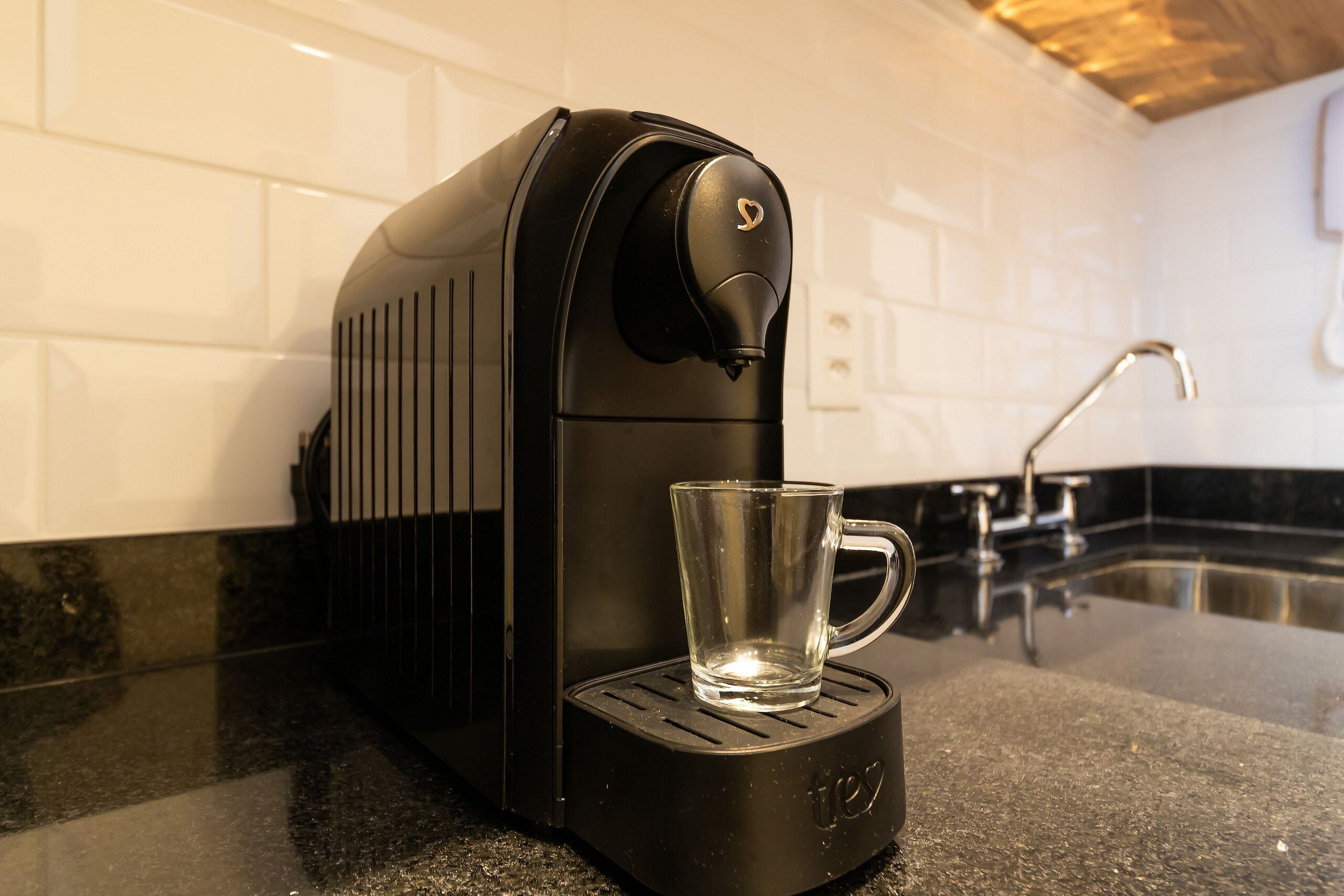 A coffee machine with a glass cup on its base, a kitchen sink, and a tiled backsplash.