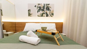 A neatly made bed with a green quilt, a wooden bedside table with a cup and a bowl, and a wall with leaf-patterned artwork.