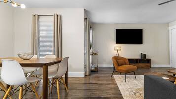 A modern living room with a dining area, wooden flooring, and a flat-screen TV mounted on the wall.