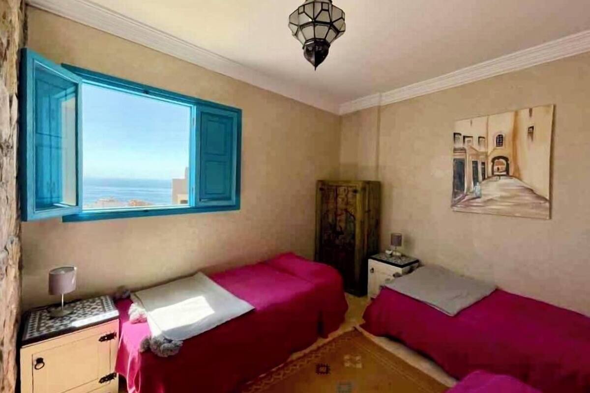 A room with two beds, a window with blue shutters, a lamp, a painting, and a small cabinet.