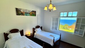 Een kamer met twee houten bedden, een kroonluchter, een schilderij en een raam met uitzicht op de oceaan.