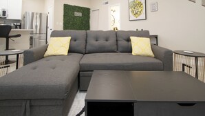 A modern living room with a grey sectional sofa, two yellow patterned pillows, a black coffee table, and a small round table with a white object on it.