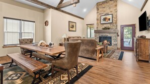 A spacious living room with a stone fireplace, wooden beams, a dining area with benches, and a television mounted on the wall.
