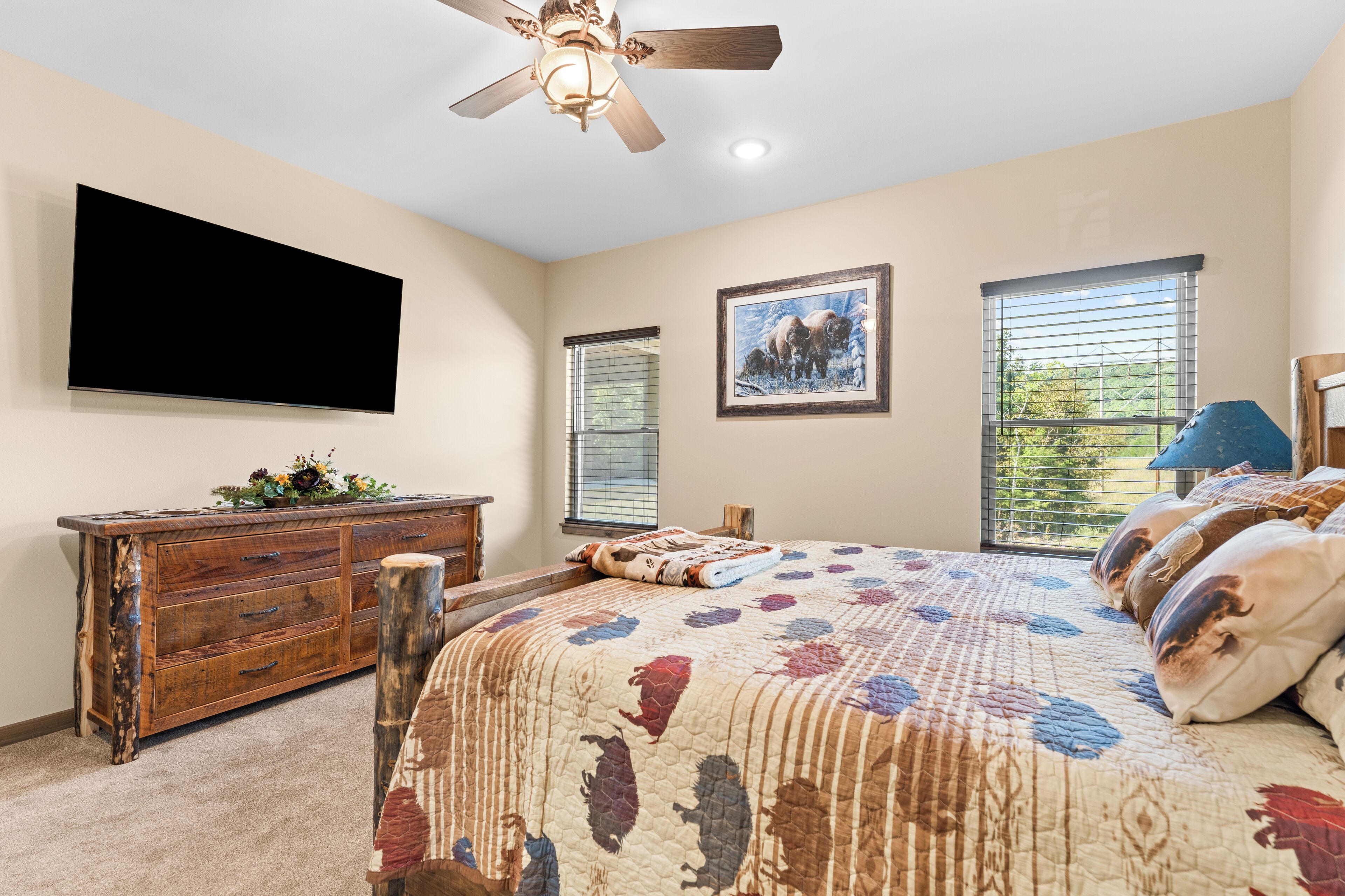 A bedroom with a bed, a wooden dresser, a ceiling fan, a flat-screen TV, and two windows with blinds.