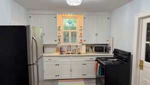 A kitchen with white cabinets, a black refrigerator, a microwave, a stove, and a window with a view of greenery. - Dog-friendly 2 bedroom beach bungalow with AC and outdoor fire-pit in Ocean View (Ocean View)