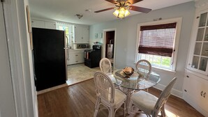 A kitchen with white cabinets, a black refrigerator, a microwave, and a stove. There is a dining table with white chairs and a glass top. A ceiling fan is visible above the table. - Dog-friendly 2 bedroom beach bungalow with AC and outdoor fire-pit in Ocean View (Ocean View)