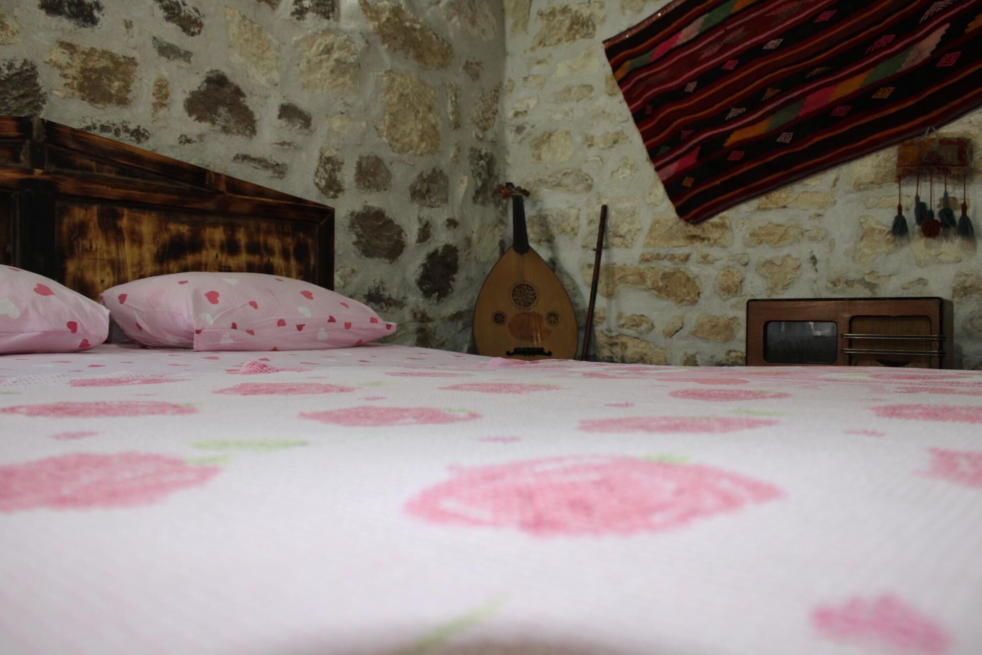 Un dormitorio con pared de piedra, una cama con colcha floral rosa, cabecero de madera, una guitarra y una radio.