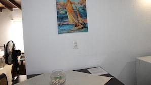 Une pièce avec un tableau représentant un voilier accroché au mur, une table sur laquelle se trouve un bocal en verre et un ventilateur dans un coin.