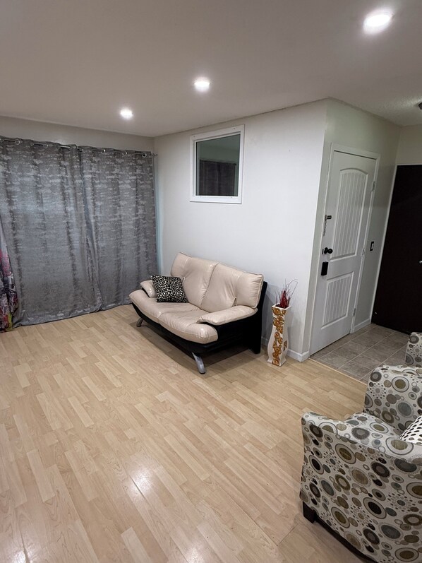 Een woonkamer met een beige bank, een gestreept fauteuil, een kleine tafel met een vaas, een deur en een raam.