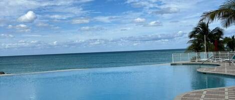A swimming pool with a tiled deck, palm trees, and a view of the ocean under a partly cloudy sky.