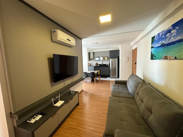 A modern living room with a flat-screen TV, a sofa, a dining area, and a kitchen in the background. - Apart. Standing on the sand in Barra Velha, 25 km from Beto Carrero (Barra Velha)