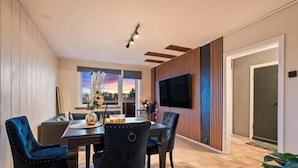 A modern dining area with a wooden floor, a dining table with chairs, a TV mounted on the wall, and a window with a view of the sunset.