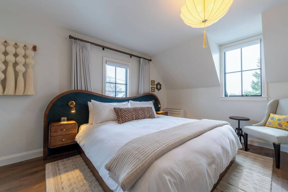 A bedroom with a large bed, a bedside table, a chair, a window, and a hanging light fixture.