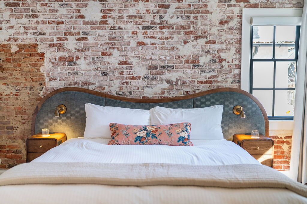 A bed with a wooden headboard, white bedding, and a floral pillow. There are two bedside tables with lamps. The room has a brick wall and a window with a view.