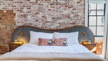 A bed with a wooden headboard, white bedding, and a floral pillow. There are two bedside tables with lamps. The room has a brick wall and a window with a view.