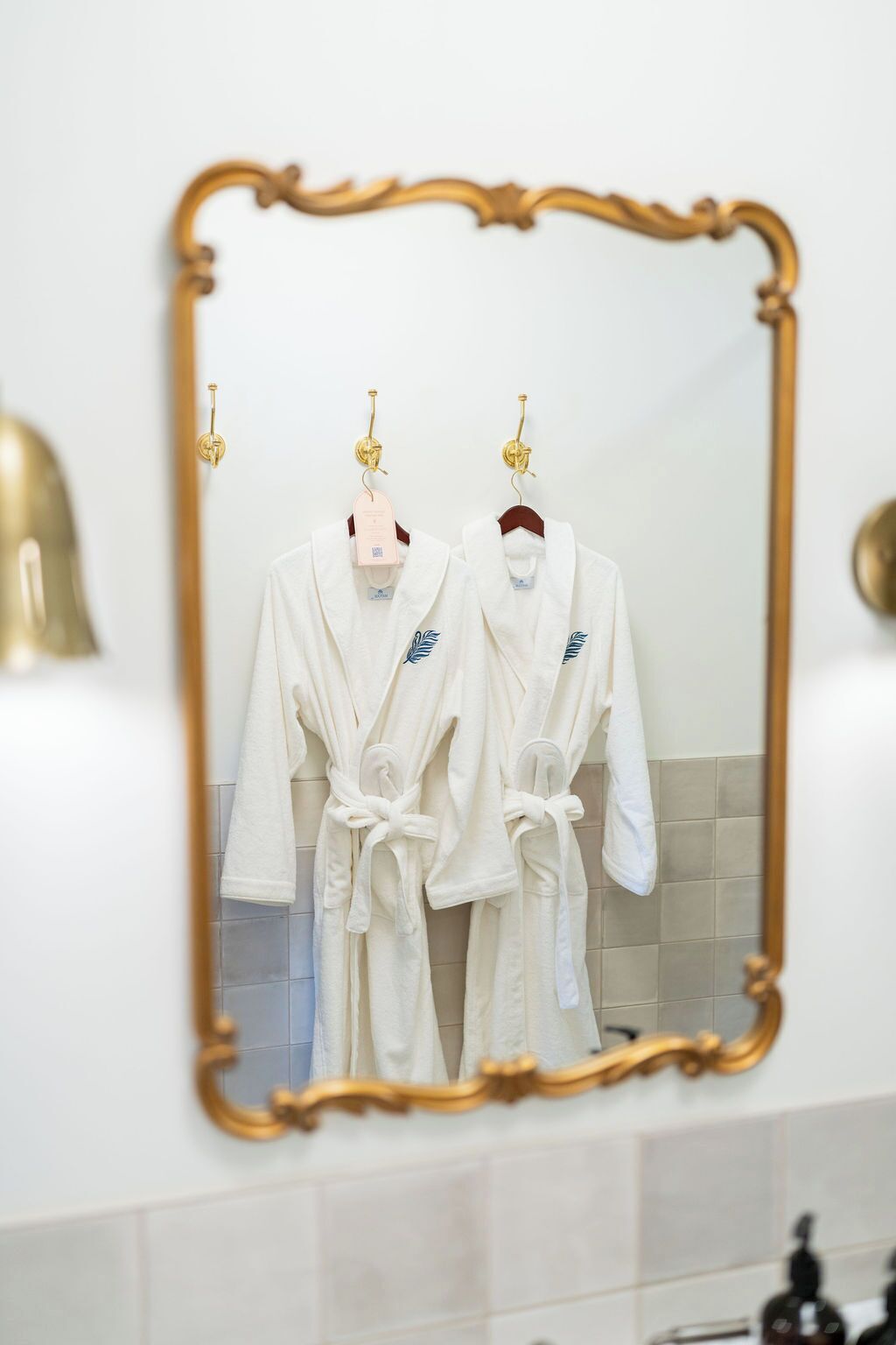 A bathroom with two white bathrobes hanging on hooks, a gold-framed mirror, and a white sink with a gold faucet.