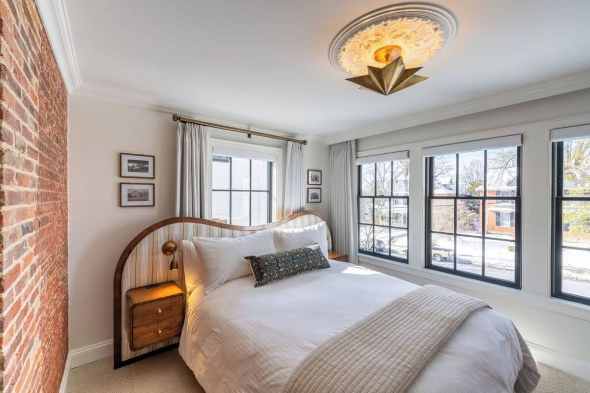 A bedroom with a large bed, a wooden nightstand, a brick wall, and large windows with curtains.