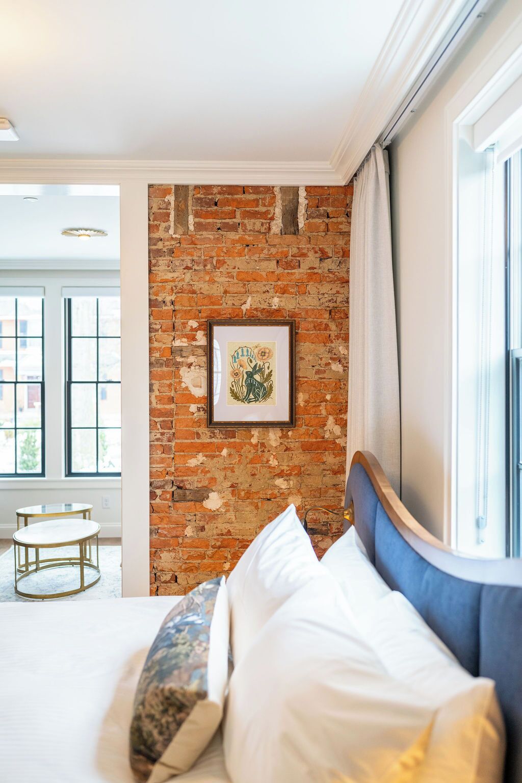 A bedroom with a brick wall, a framed picture, a bed with white and blue bedding, and a window with curtains.