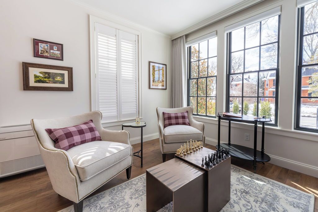 Una habitación con dos sillones, una mesa de ajedrez y ventanas amplias con persianas.