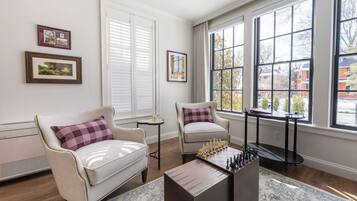 Una habitación con dos sillones, una mesa de ajedrez y ventanas amplias con persianas.