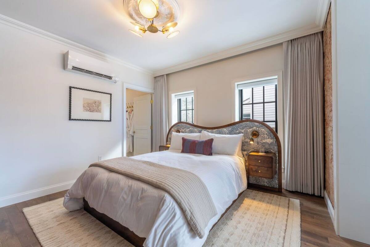 A bedroom with a large bed, a nightstand, an air conditioning unit, a framed picture on the wall, and a window with curtains.