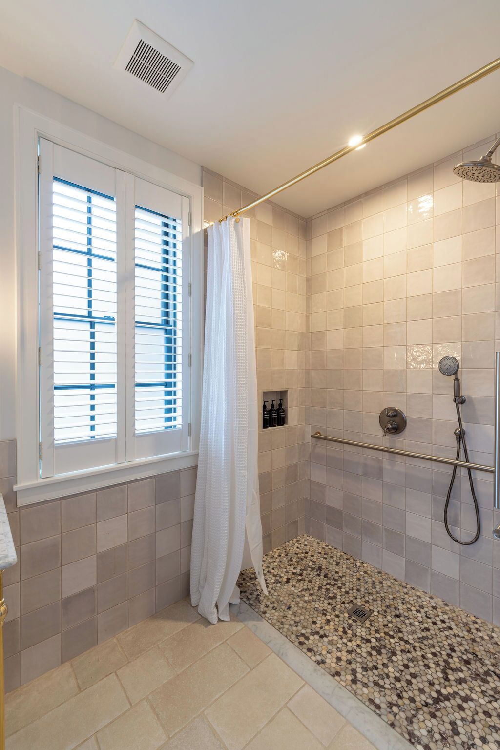 A shower with a handheld showerhead, a window with blinds, and a tiled floor.