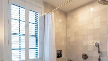 A shower with a handheld showerhead, a window with blinds, and a tiled floor.