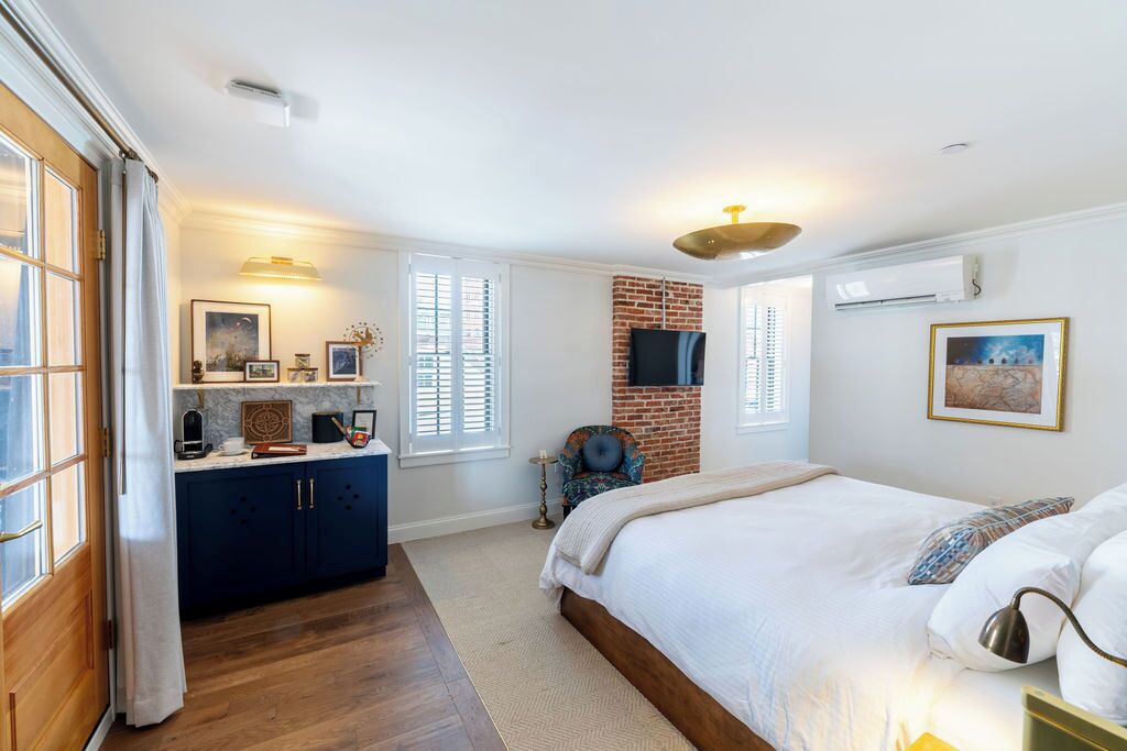 A bedroom with a large bed, a brick accent wall, a TV mounted on the wall, and a window with blinds.