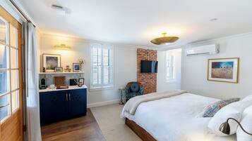 A bedroom with a large bed, a brick accent wall, a TV mounted on the wall, and a window with blinds.