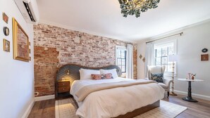 A bedroom with a brick wall, a large bed with white bedding, a side table, a chair, and a small round table with a lamp. - The Francis Hotel & Suites (Kennett Square)