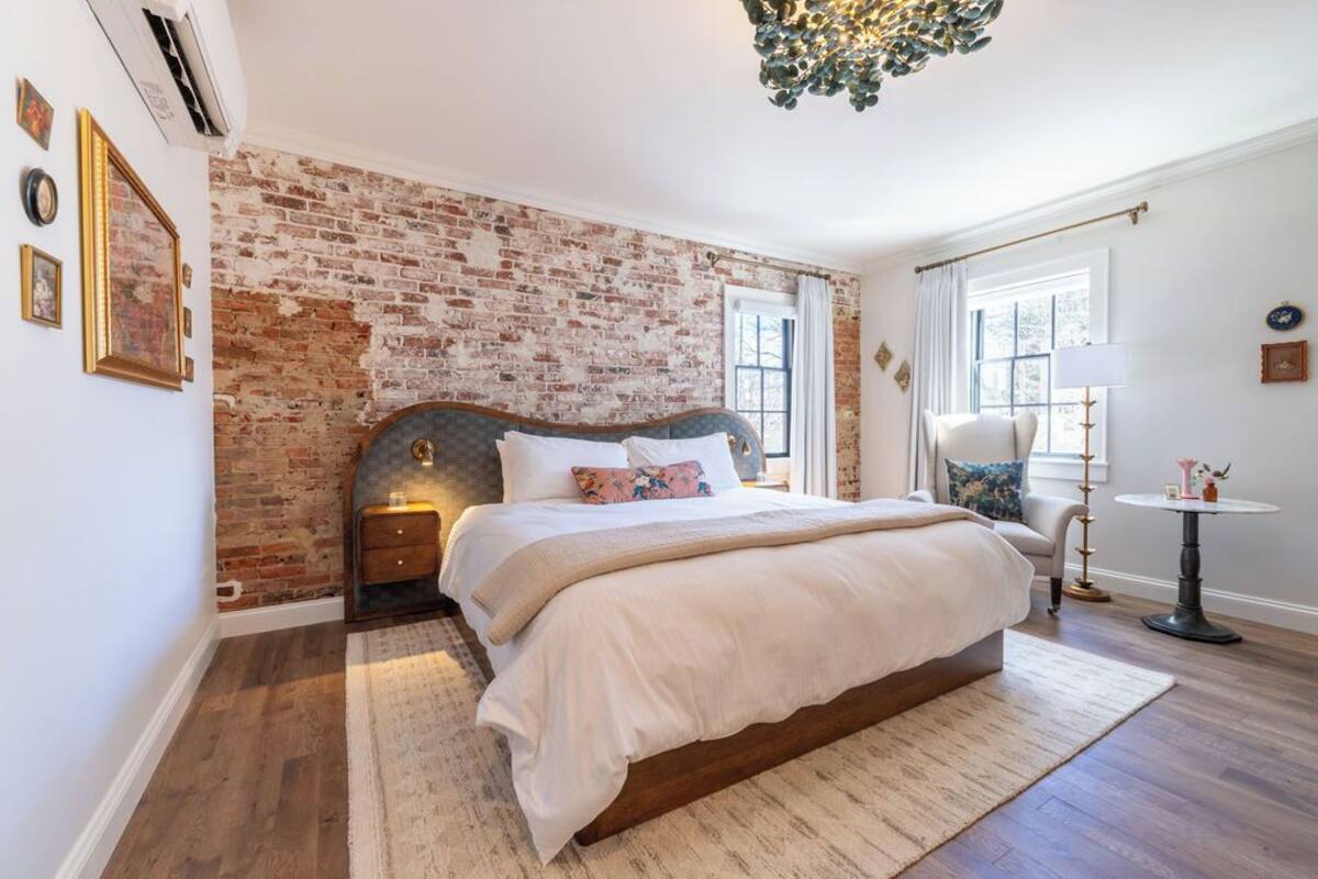 A bedroom with a brick wall, a large bed with white bedding, a side table, a chair, and a small round table with a lamp.