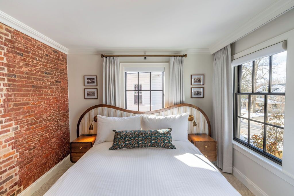 Un dormitorio con pared de ladrillo, un ventanal con cortinas, estructura de cama de madera y mesita de noche con lámpara.