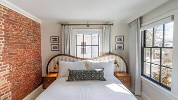 Un dormitorio con pared de ladrillo, un ventanal con cortinas, estructura de cama de madera y mesita de noche con lámpara.