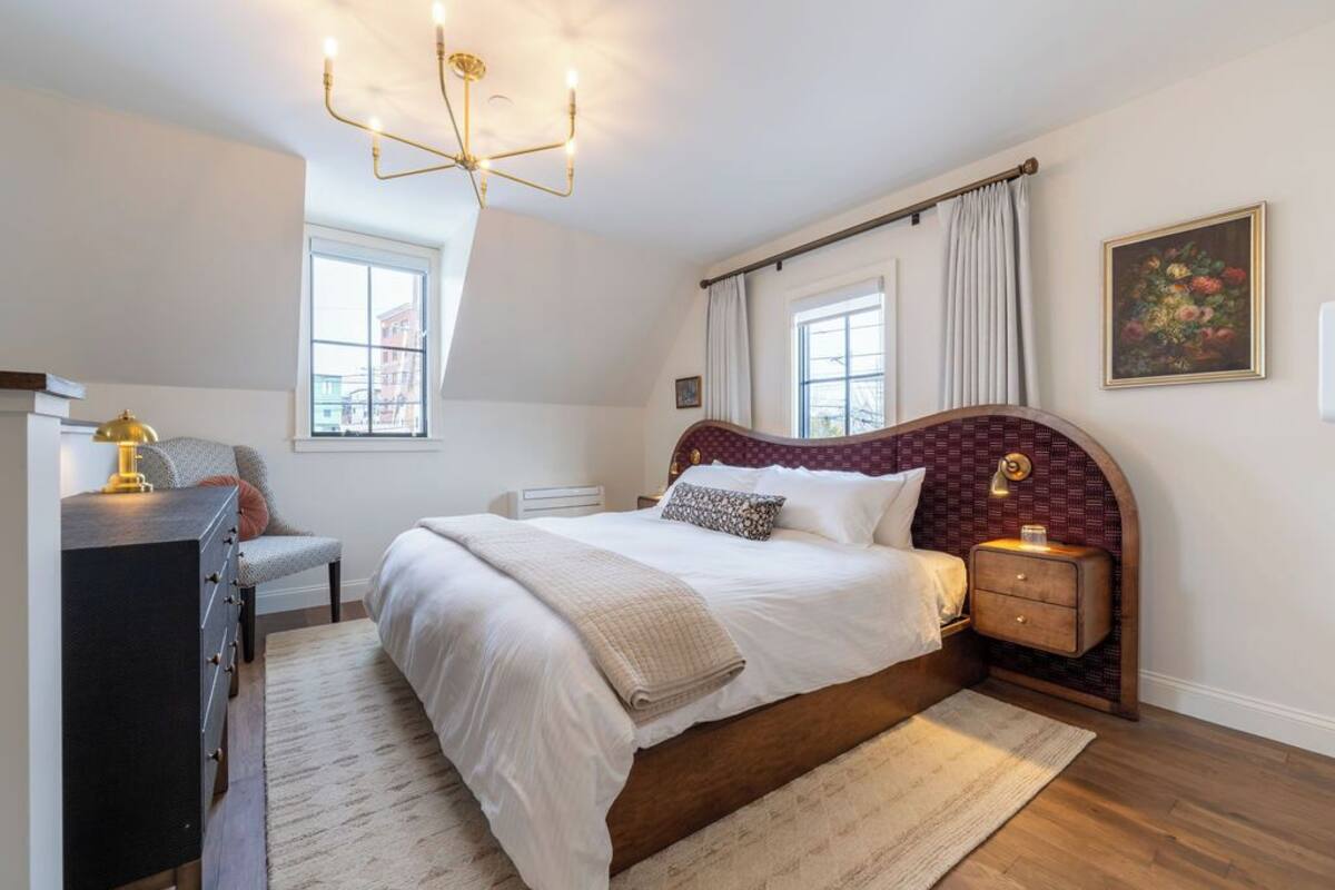 A bedroom with a large bed, a dresser, a nightstand, a chandelier, and a window with curtains.