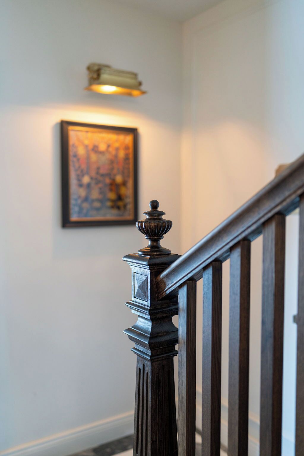 Un escalier avec une main courante et des balustres en bois foncé, menant à un palier où se trouve un luminaire fixé au mur et une œuvre d’art encadrée.