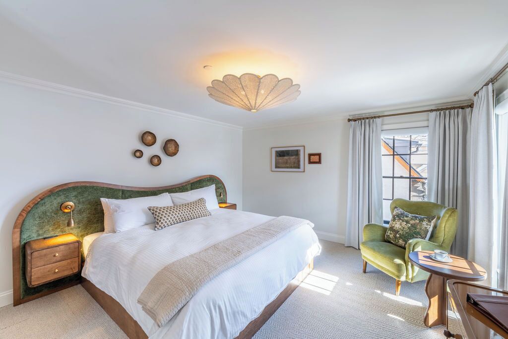 Ein helles Schlafzimmer mit einem großen Bett, einem grünen Sessel, einem kleinen Tisch und einer dekorativen Deckenleuchte.