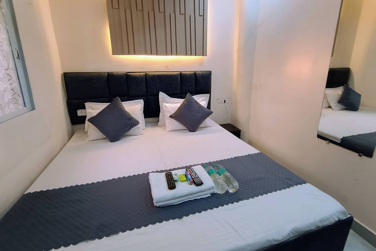 A hotel room with a bed, two pillows, a bedside table, a mirror, and a wall-mounted light fixture.