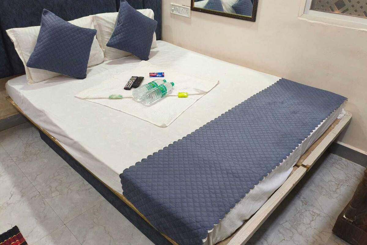 A neatly made bed with a white mattress, two pillows, a blue quilted blanket, and a small table with a remote control, water bottle, and tissues.