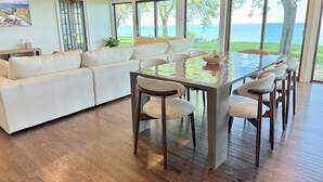Una sala de estar moderna con un sofá grande, una mesa de comedor con sillas y ventanas amplias que dan a una vista del mar.