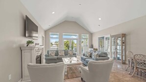 A spacious living room with a high ceiling, a fireplace, a large TV, and a dining area with wicker chairs.
