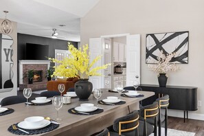 Un coin repas avec une longue table dressée pour six personnes, une cheminée et, au fond, une cuisine moderne.