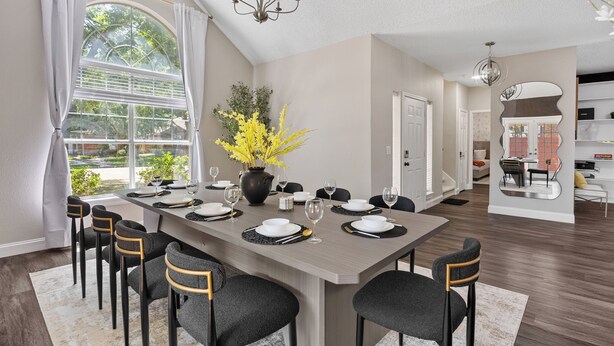 A dining area with a large table set for six, surrounded by chairs, a vase of yellow flowers, and a view of greenery through a large window. - The Pony Haus – Pool, Spa +Climbing Wall sleeps 11 in Frisco TX! (Frisco)