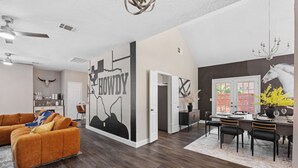 A modern living room with a sectional sofa, a dining area with a table set for four, and a kitchen in the background. - The Pony Haus – Pool, Spa +Climbing Wall sleeps 11 in Frisco TX! (Frisco)