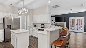 A modern kitchen with white cabinetry, stainless steel appliances, a central island with bar stools, and a dining area with a black accent wall. - The Pony Haus – Pool, Spa +Climbing Wall sleeps 11 in Frisco TX! (Frisco)
