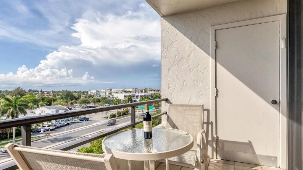 Un balcon avec une table en verre, une chaise et une bouteille de vin. Le balcon donne sur une rue où l’on peut voir des voitures et des immeubles.