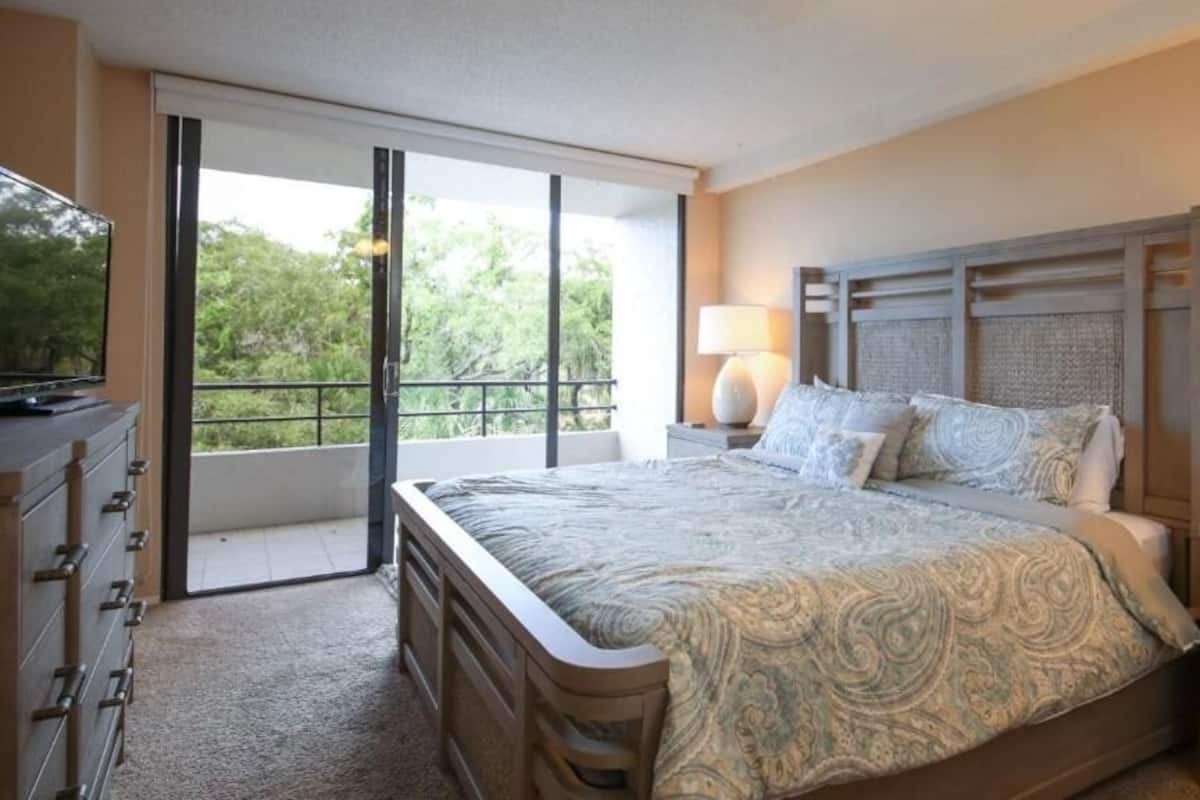 A bedroom with a large bed, a dresser, a nightstand with a lamp, and a sliding glass door leading to a balcony.