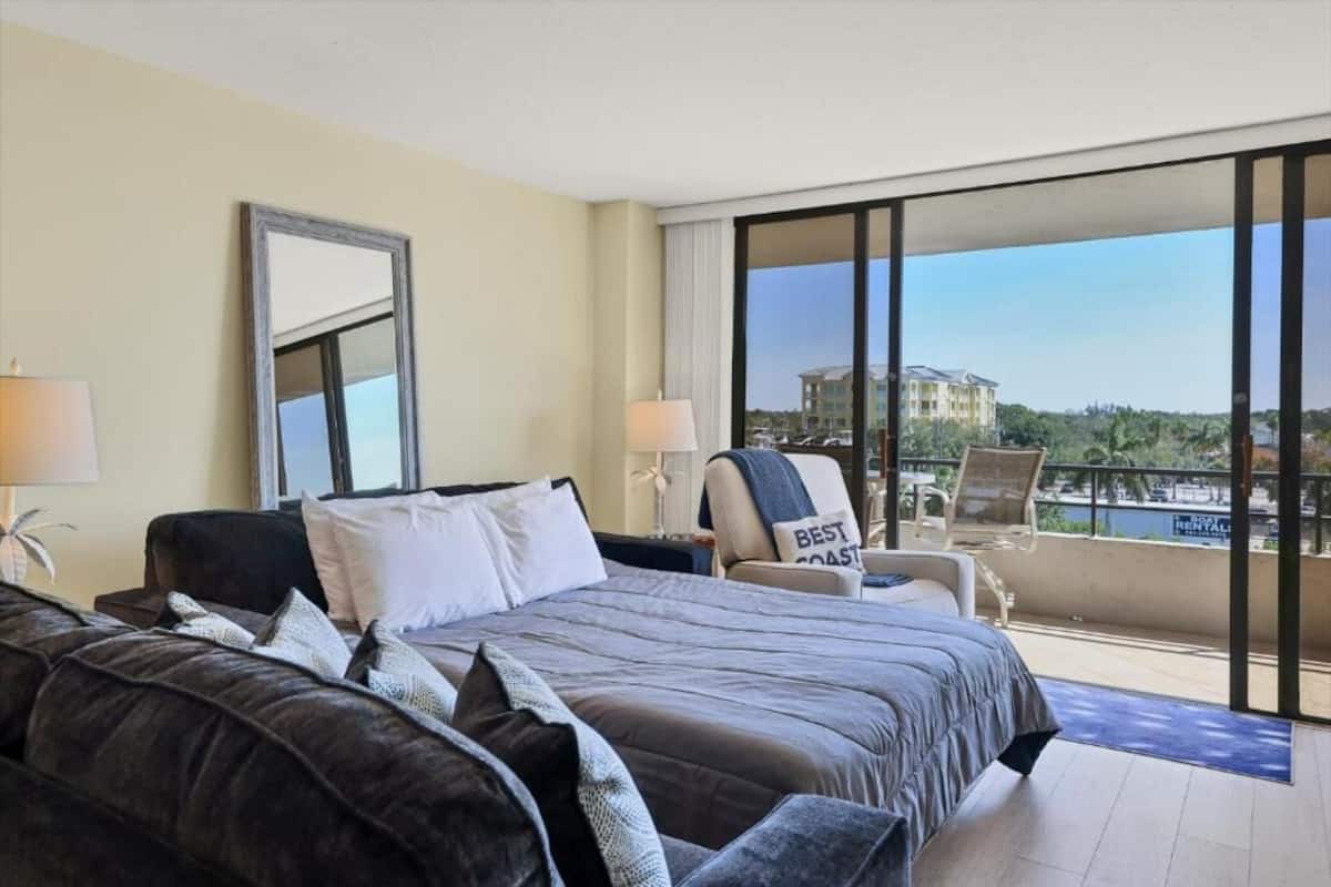A hotel room with a large bed, a chair with a 'Best Coast' sign, a balcony with a view of buildings and greenery.